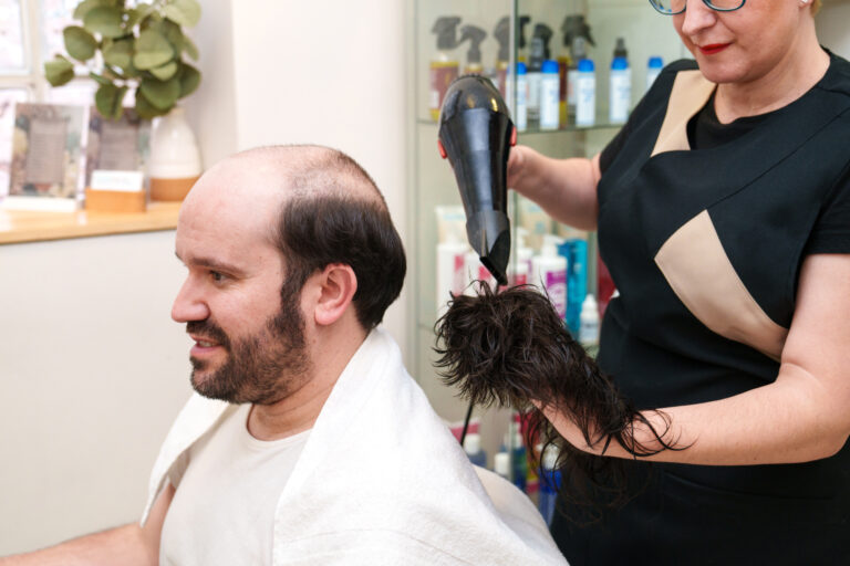 A hairdresser uses a hairdryer with a wig attachment to expertly style a smiling man's hair, creating a look that perfectly complements wigs and hairpieces.