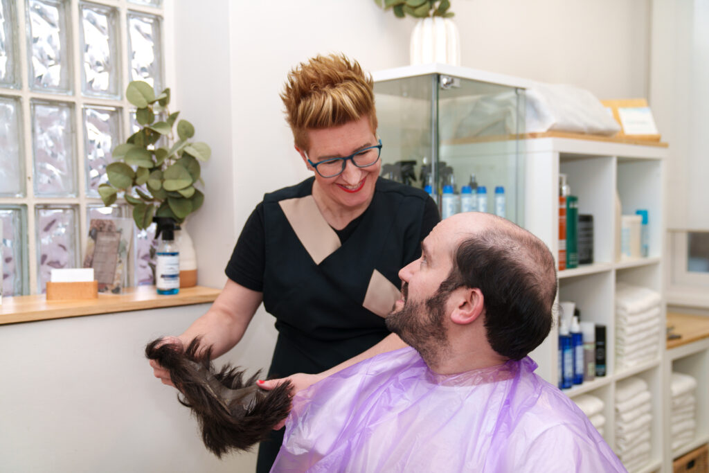 A barber, smiling confidently, holds a hairpiece as the man in the purple cape sits in the salon chair, hopeful for that perfect hairline.