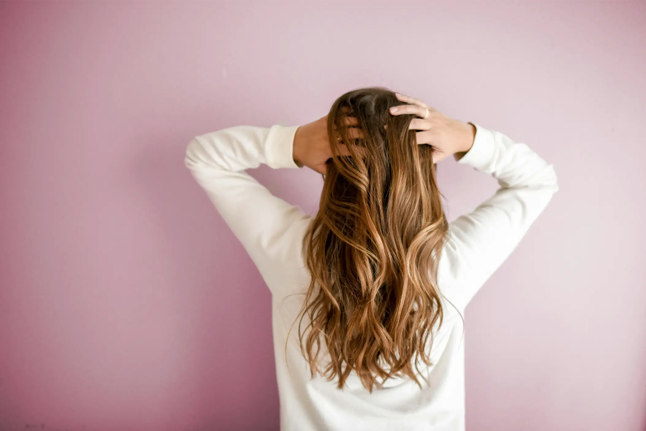 A person with long, wavy hair touches their head in contemplation, facing away from the camera against a light pink background. Their serene pose invites thoughts about what can make your hair fall out despite its apparent healthy state. Reading this blog will help you understand what can make your hair fall out